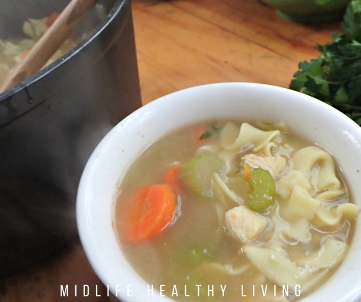 A pot with wooden ladle and a white small bowl both filled with homemade chicken noodle soup.