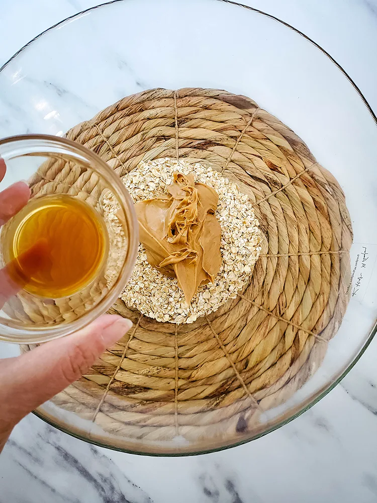 Overhead view of oats, peanut butter in a clear mixing bowl with vanilla extract in a small bowl in the forefront.