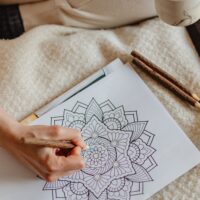 Person sitting down coloring in a mandala illustration while holding a cup of coffee and relaxing