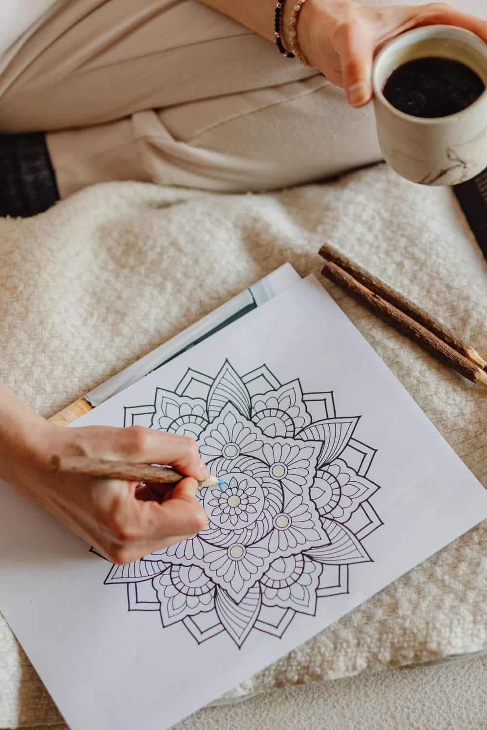 Person sitting down coloring in a mandala illustration while holding a cup of coffee and relaxing